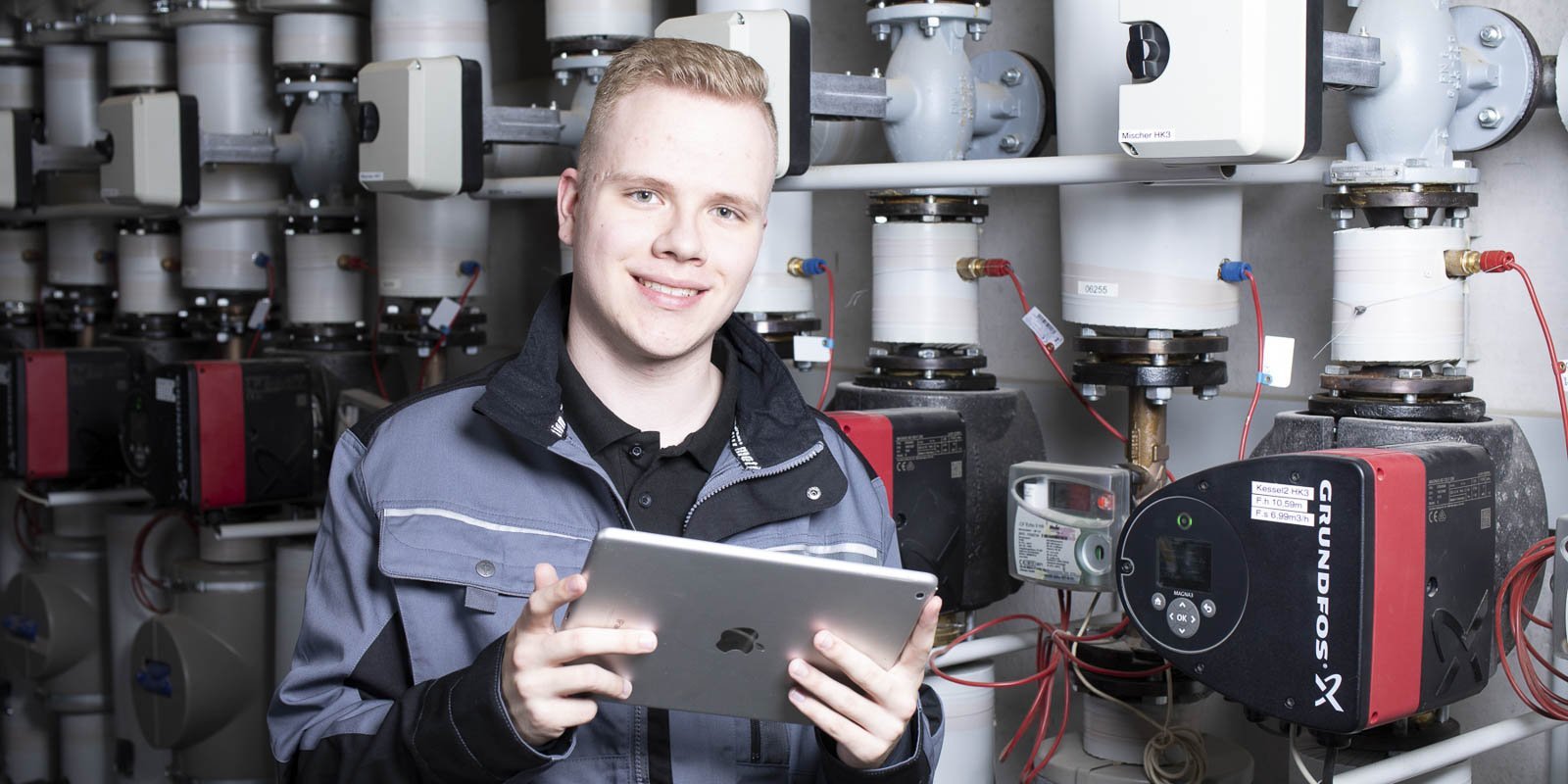 Ein junger Anlagenmechaniker mit einem Tablet in der Hand vor einer Heizungsanlage