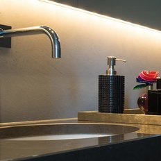 Der glänzende Wasserhahn über einem runden Waschbecken, umgeben von dekorativen Flaschen und einer bunten Blume.