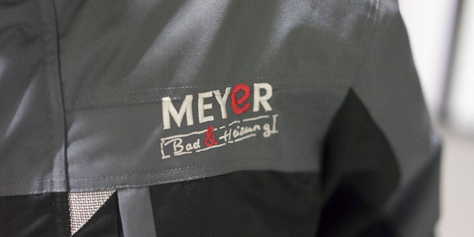 Die Jacke zeigt das MEYER-Logo in rot und weiß, das auf einem grauen Stoff mit schwarzem Unterteil platziert ist.